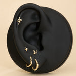 A black mannequin ear displays five gold earrings, including a 14K Solid Yellow Gold Snake Flat Back Stud at the top, alongside a small gold hoop, textured hoop, star stud, and bird earring, all set against a beige background.