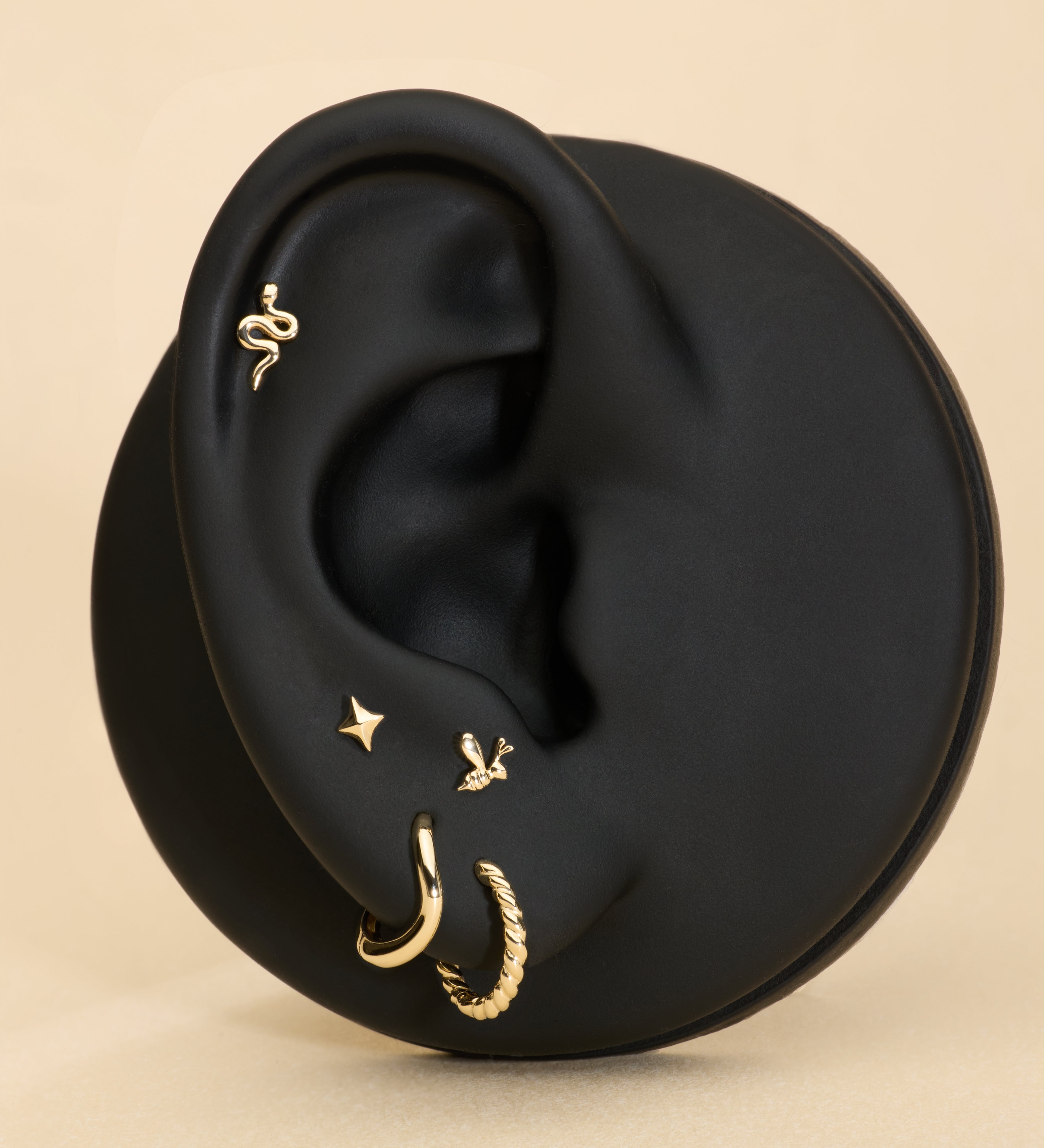 A black mannequin ear displays five gold earrings, including a 14K Solid Yellow Gold Snake Flat Back Stud at the top, alongside a small gold hoop, textured hoop, star stud, and bird earring, all set against a beige background.
