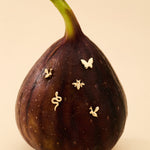 A ripe fig stands upright against a beige background, adorned with the 14K Solid White Gold Bumble Bee Flat Back Stud (Left) alongside other delicate gold pins.