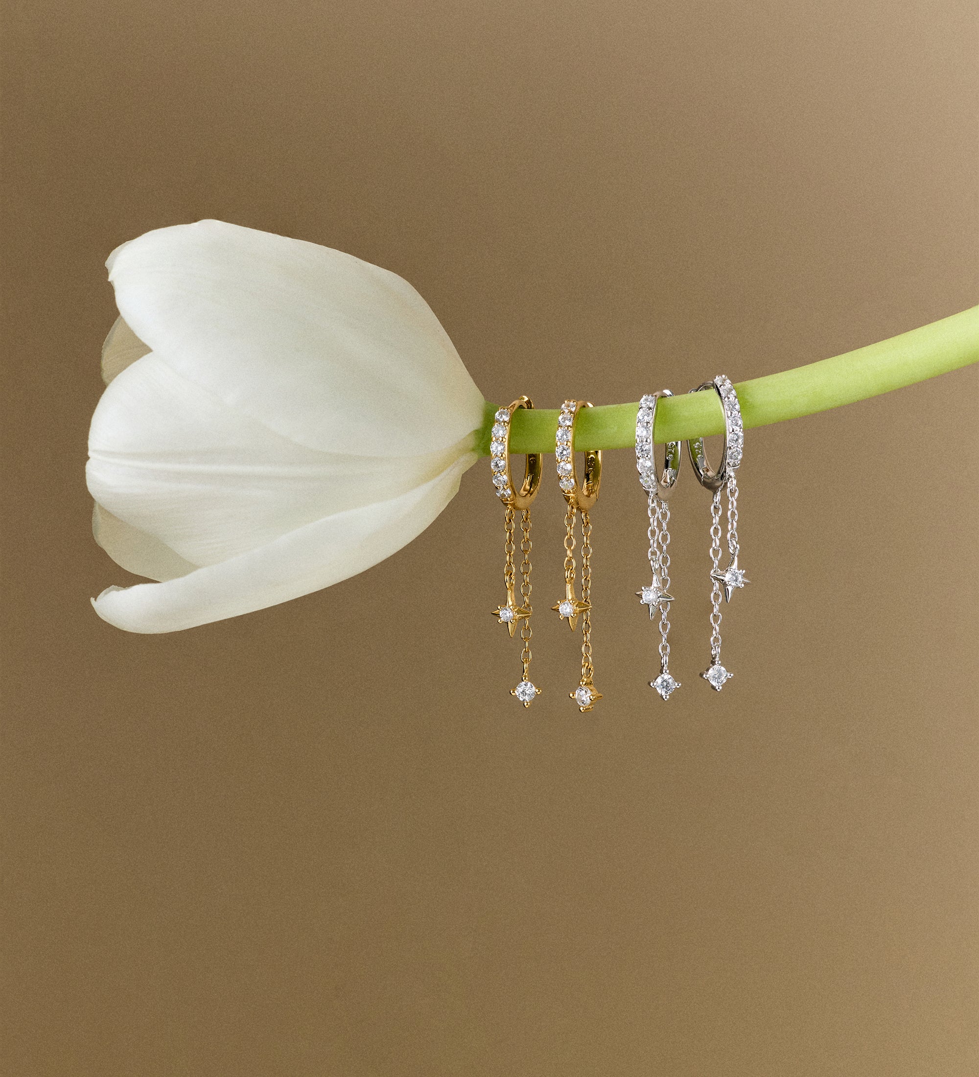 A white tulip with four pairs of earrings—including Star Double Chain Dangle Huggies in Gold—dangling from its stem, set against a plain beige background.