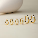 Six pairs of Nori Twisted Hoop Earrings in Gold, including one with embedded crystals, are arranged on a neutral beige surface. A smooth, white, egg-shaped object appears blurred in the background.