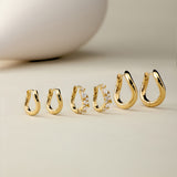 Six pairs of Nori Twisted Hoop Earrings in Gold, including one with embedded crystals, are arranged on a neutral beige surface. A smooth, white, egg-shaped object appears blurred in the background.