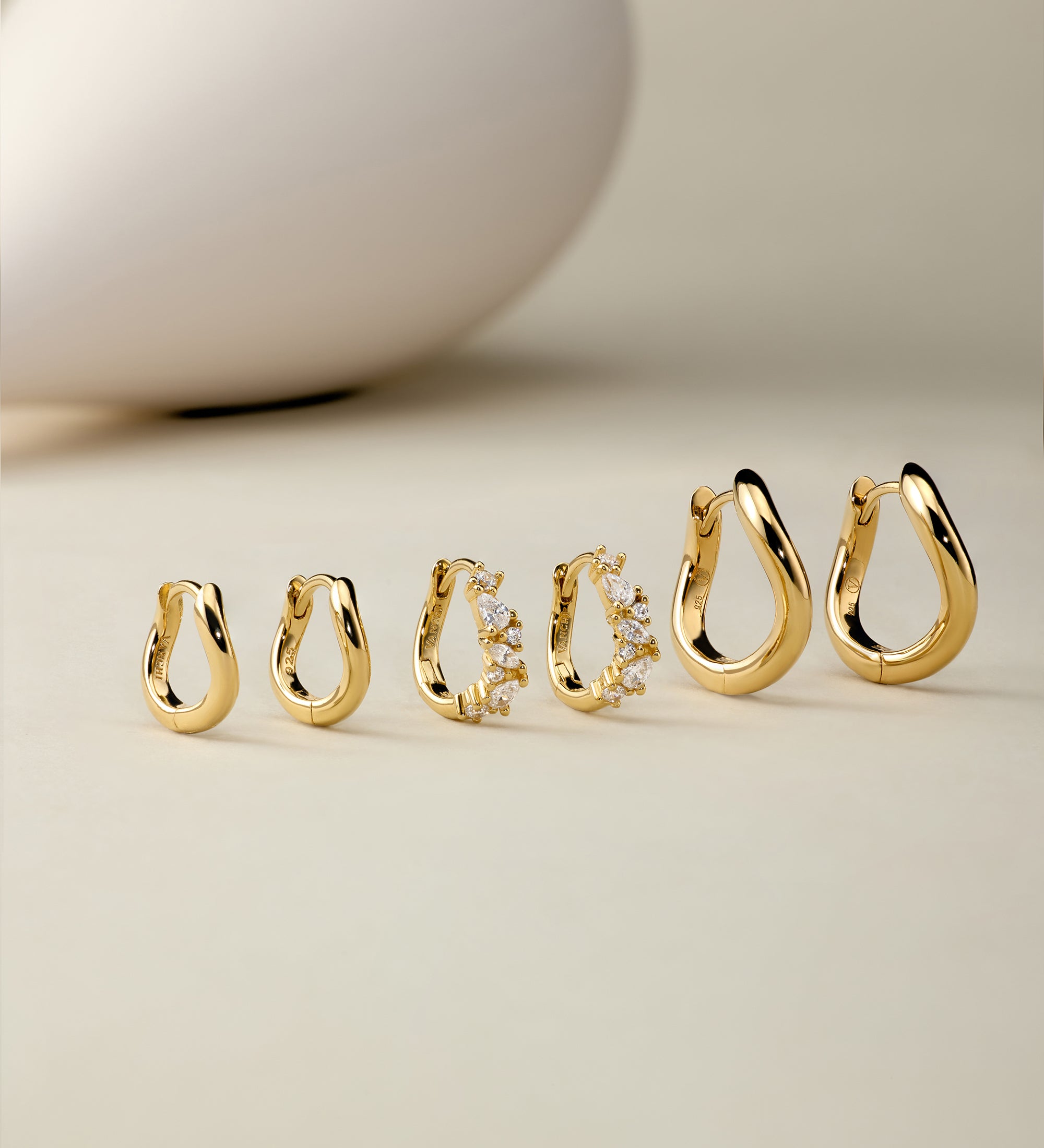 Six pairs of Nori Twisted Hoop Earrings in Gold, including one with embedded crystals, are arranged on a neutral beige surface. A smooth, white, egg-shaped object appears blurred in the background.