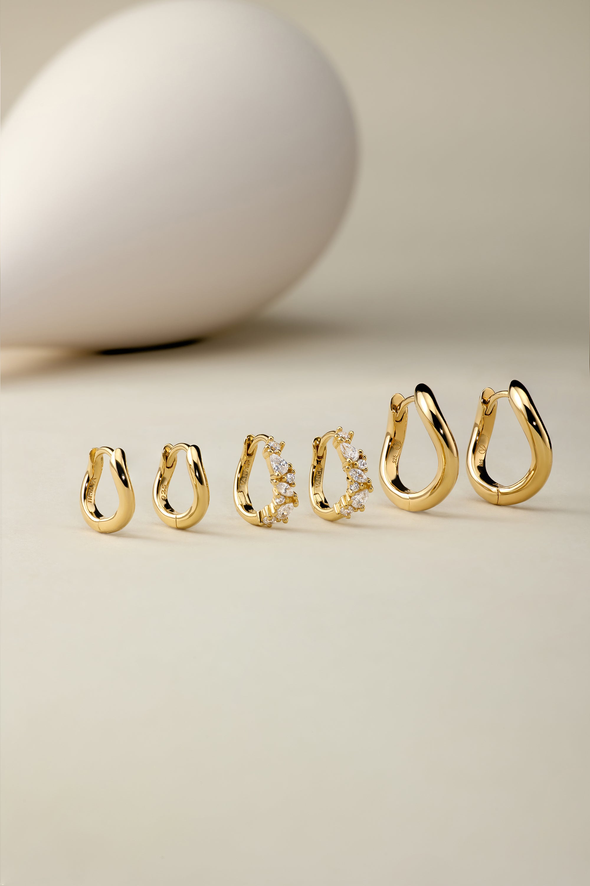 Six pairs of Nori Twisted Hoop Earrings in Gold, including one with embedded crystals, are arranged on a neutral beige surface. A smooth, white, egg-shaped object appears blurred in the background.