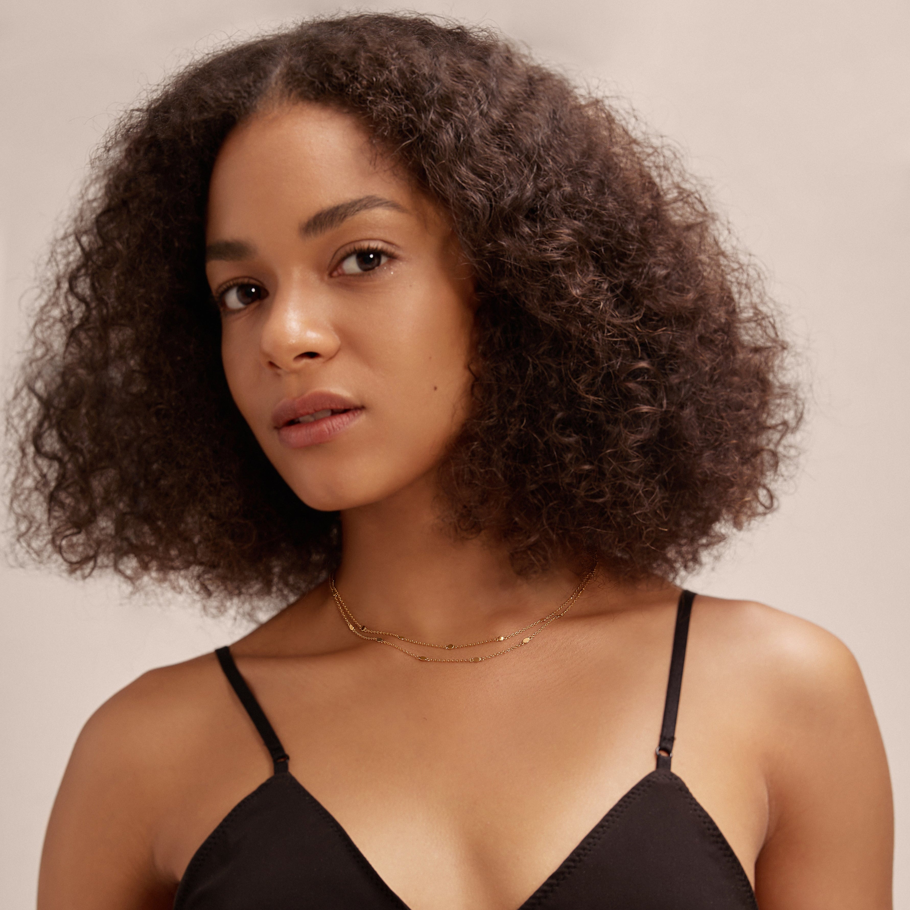 A woman with curly hair stands against a neutral background, wearing a black strappy top and delicate gold jewelry, including rings, bracelets, and the Simona Double Chain Stacking Necklace in Gold.