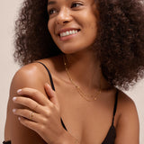A woman with curly hair in a black tank top smiles to the side, showcasing the Simona Double Chain Stacking Necklace in Gold along with a gold bracelet and rings, set against a plain light background.