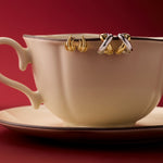 A cream teacup and saucer sit on a red surface, with two pairs of Mia Two-tone Mixed Metal Hoops hanging from the rim, set against a deep red background—an eye-catching way to display versatile jewellery.