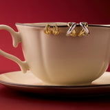 A cream teacup and saucer sit on a red surface, with two pairs of Mia Two-tone Mixed Metal Hoops hanging from the rim, set against a deep red background—an eye-catching way to display versatile jewellery.