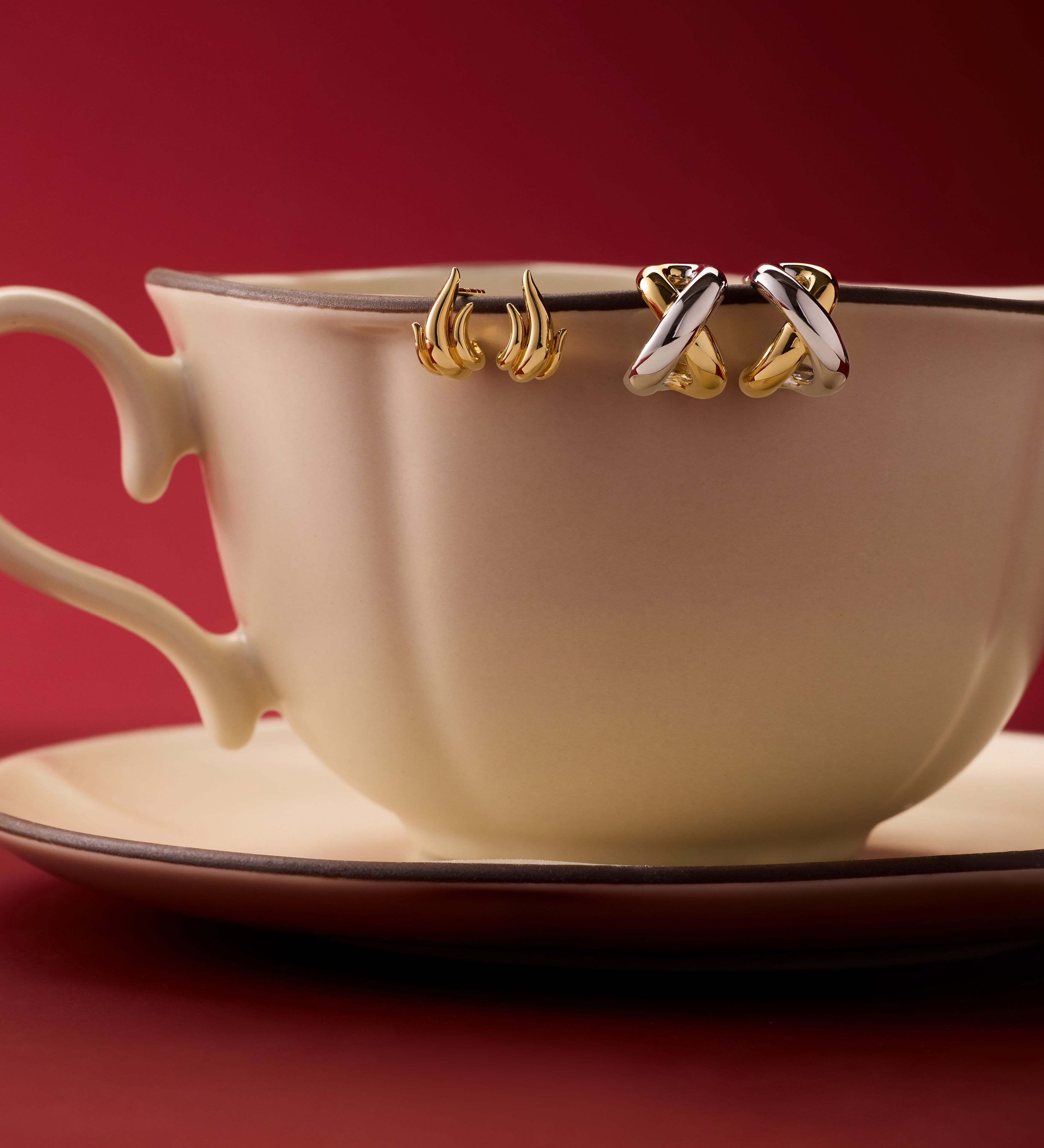 A cream teacup and saucer sit on a red surface, with two pairs of Mia Two-tone Mixed Metal Hoops hanging from the rim, set against a deep red background—an eye-catching way to display versatile jewellery.