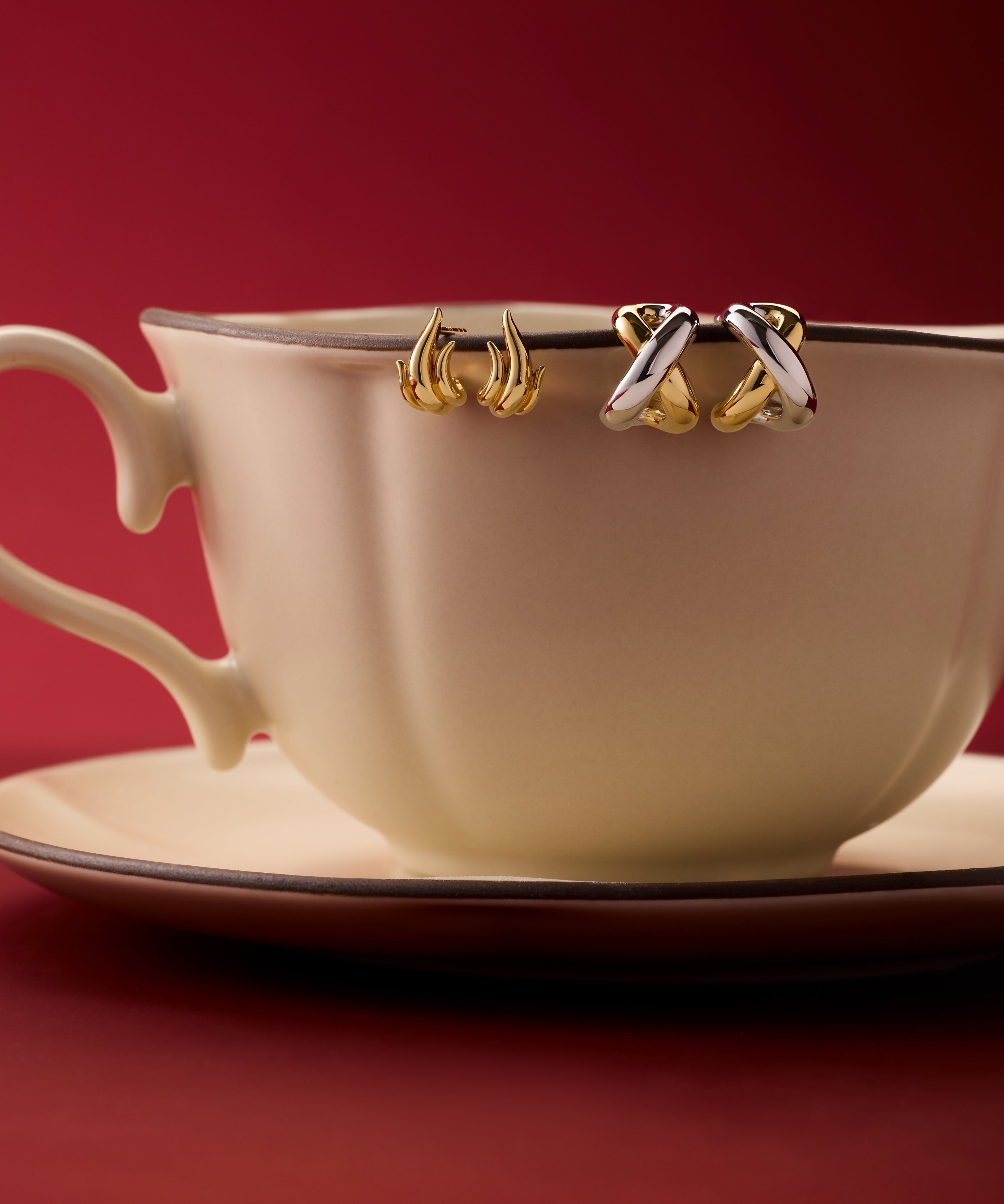 A cream teacup and saucer sit on a red surface, with two pairs of Mia Two-tone Mixed Metal Hoops hanging from the rim, set against a deep red background—an eye-catching way to display versatile jewellery.