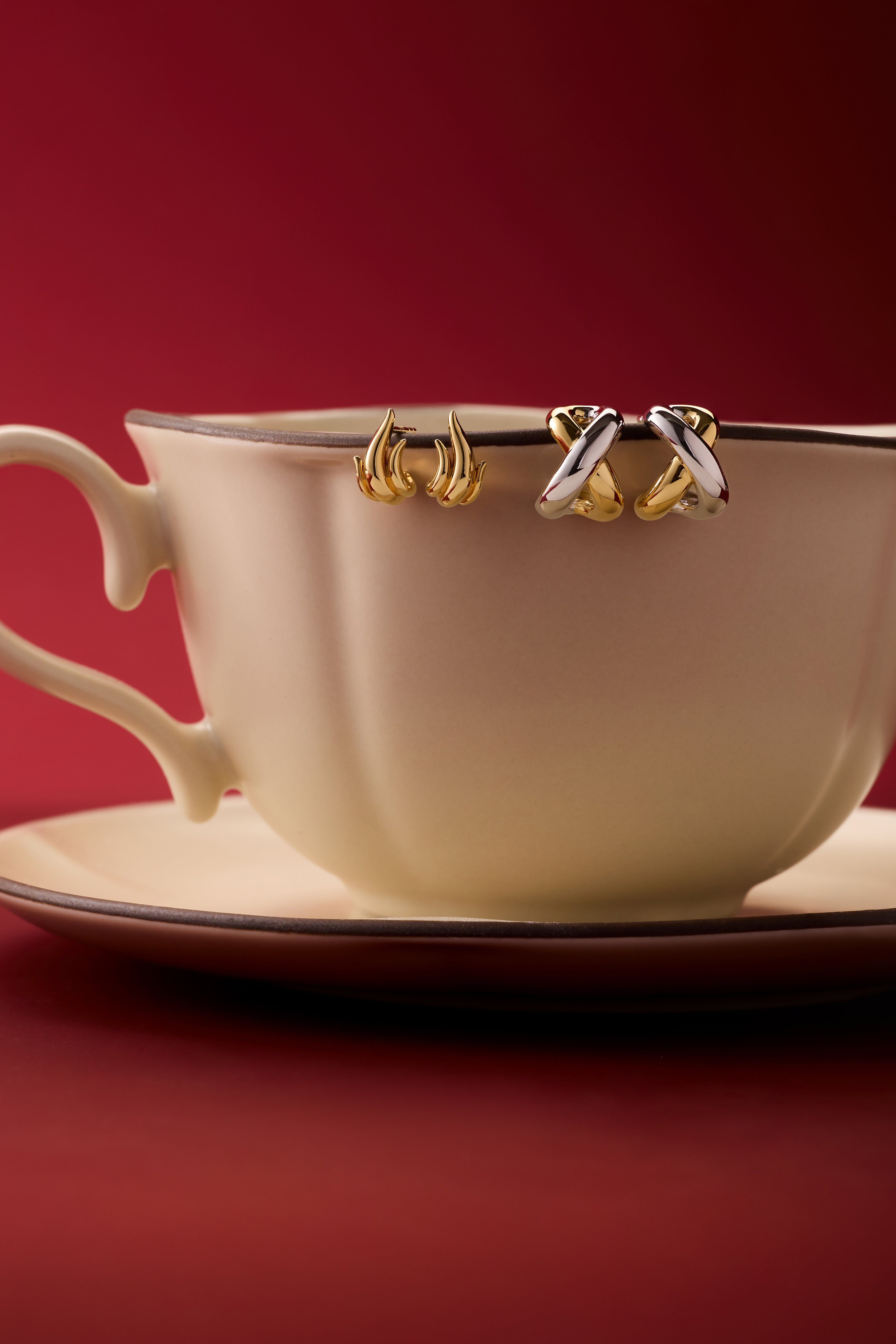 A cream teacup and saucer sit on a red surface, with two pairs of Mia Two-tone Mixed Metal Hoops hanging from the rim, set against a deep red background—an eye-catching way to display versatile jewellery.