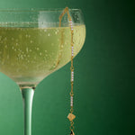 A coupe glass of sparkling wine sits against a green background, with the Pia Crystal Link Bracelet in Gold elegantly draped over the rim as bubbles glimmer in the beverage.