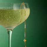 A coupe glass of sparkling wine sits against a green background, with the Pia Crystal Link Bracelet in Gold elegantly draped over the rim as bubbles glimmer in the beverage.