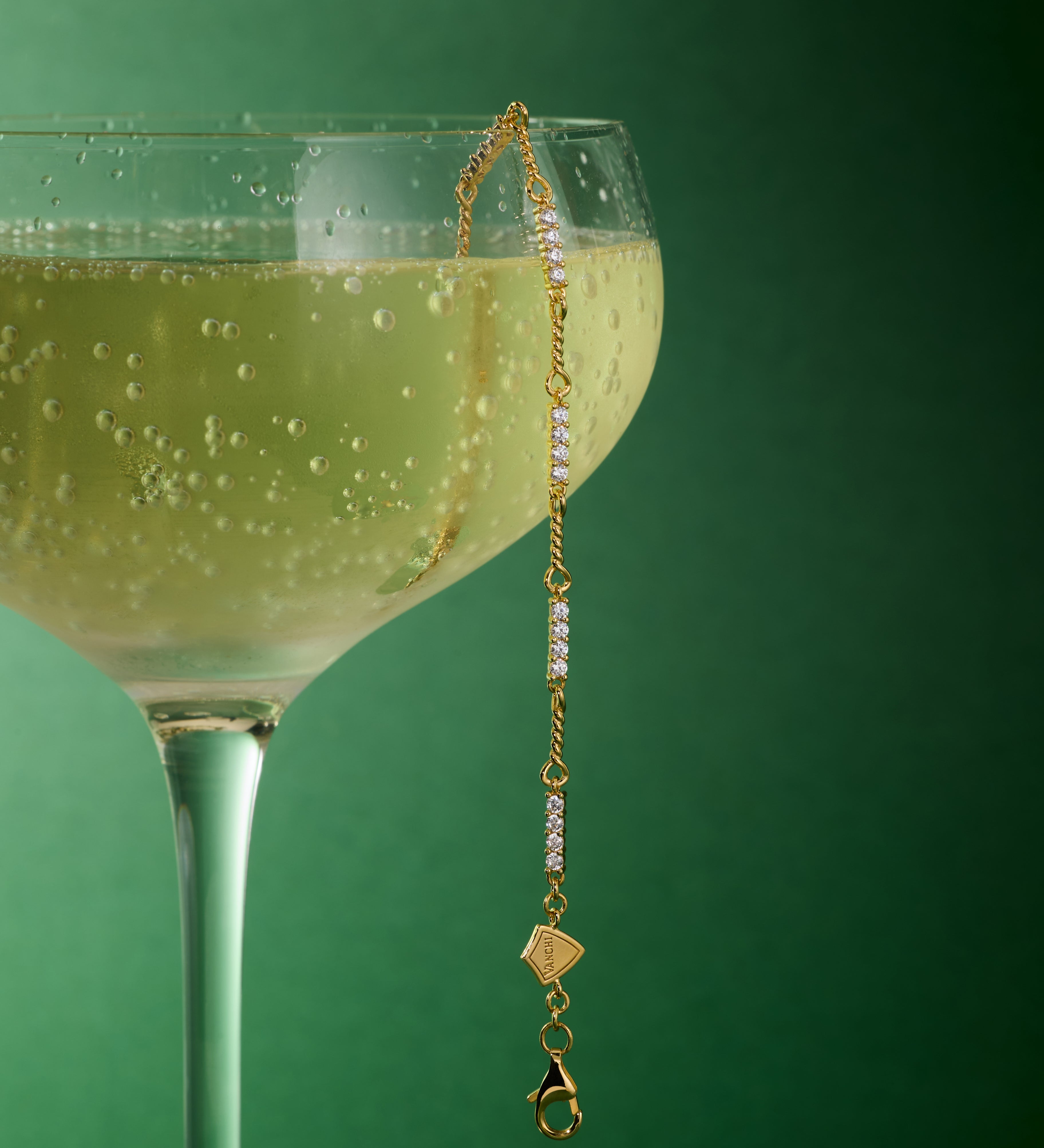 A coupe glass of sparkling wine sits against a green background, with the Pia Crystal Link Bracelet in Gold elegantly draped over the rim as bubbles glimmer in the beverage.