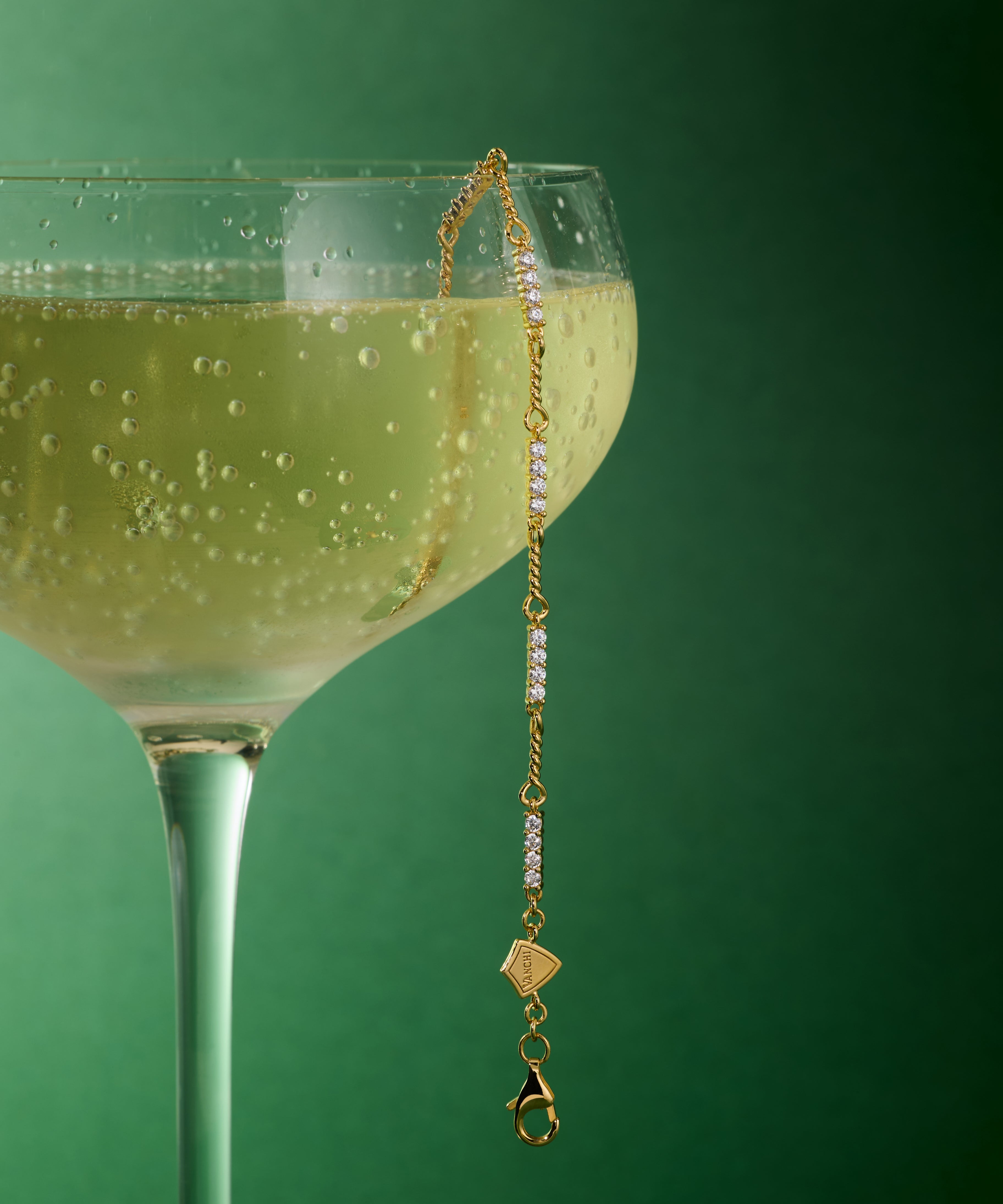 A coupe glass of sparkling wine sits against a green background, with the Pia Crystal Link Bracelet in Gold elegantly draped over the rim as bubbles glimmer in the beverage.