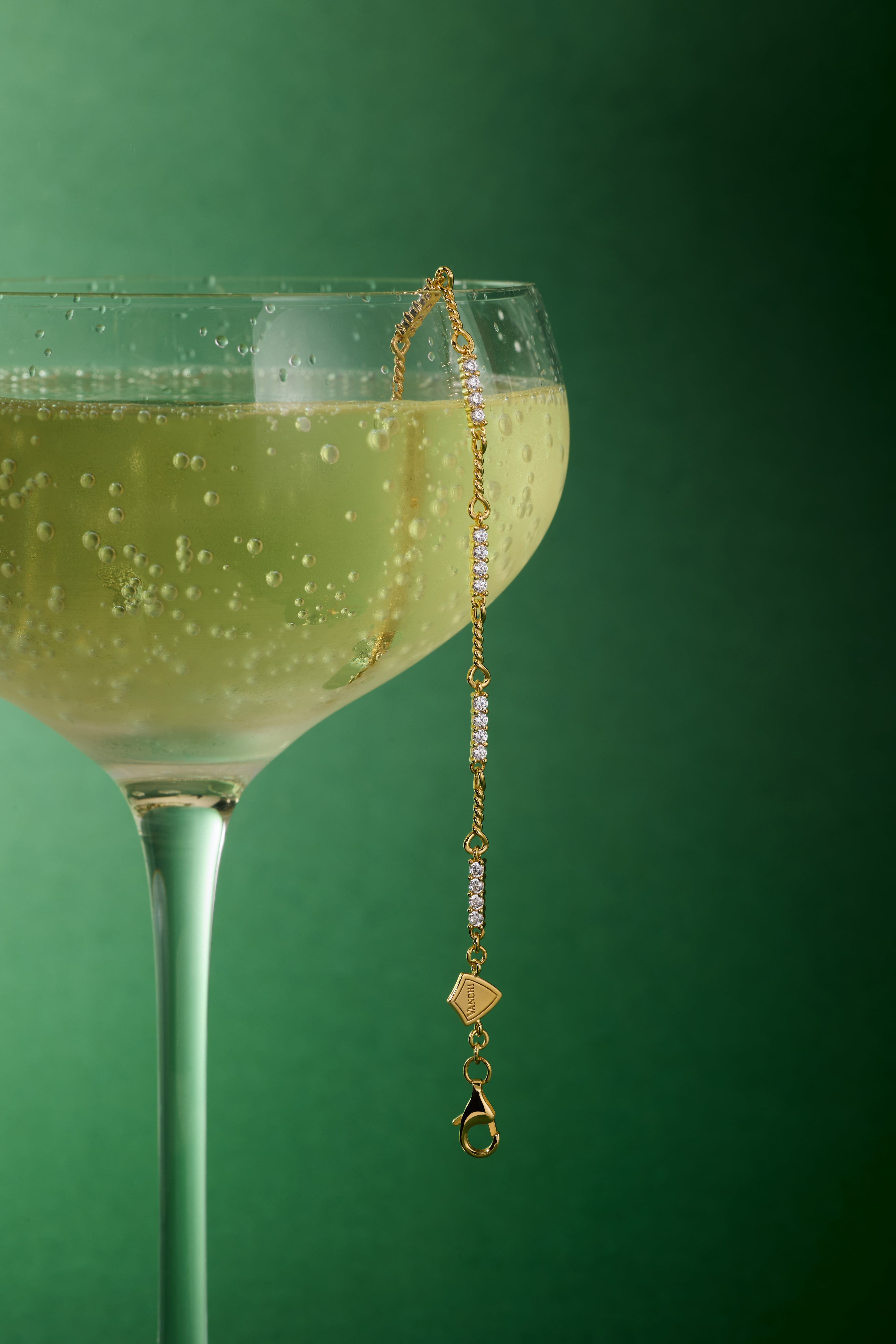 A coupe glass of sparkling wine sits against a green background, with the Pia Crystal Link Bracelet in Gold elegantly draped over the rim as bubbles glimmer in the beverage.