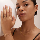 A woman with smooth, medium skin tone wears layered gold jewelry, including the Simona Double Chain Stacking Necklace in Gold. Posing with one hand on a white sphere, she gazes confidently at the camera in a black top and neatly styled hair.