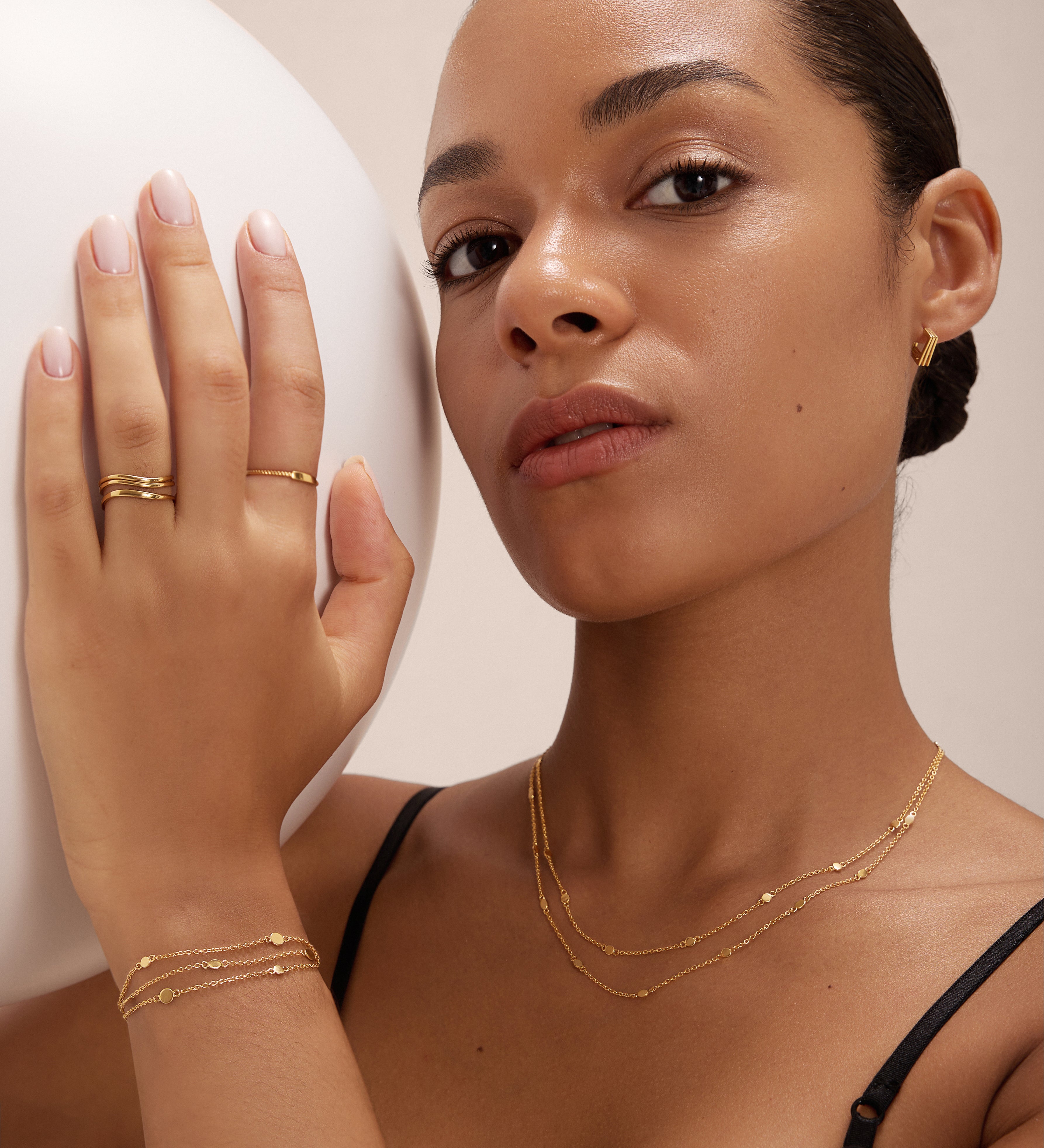A woman with smooth, medium skin tone wears layered gold jewelry, including the Simona Double Chain Stacking Necklace in Gold. Posing with one hand on a white sphere, she gazes confidently at the camera in a black top and neatly styled hair.
