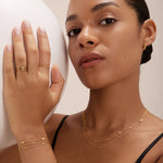 A woman with smooth skin and tied-back hair wears a black top, gold rings, small hoop earrings, and the Simone Triple Chain Layered Bracelet in Gold. She holds a large white sphere in her left hand.