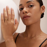 A woman with smooth skin and tied-back hair wears a black top, gold rings, small hoop earrings, and the Simone Triple Chain Layered Bracelet in Gold. She holds a large white sphere in her left hand.