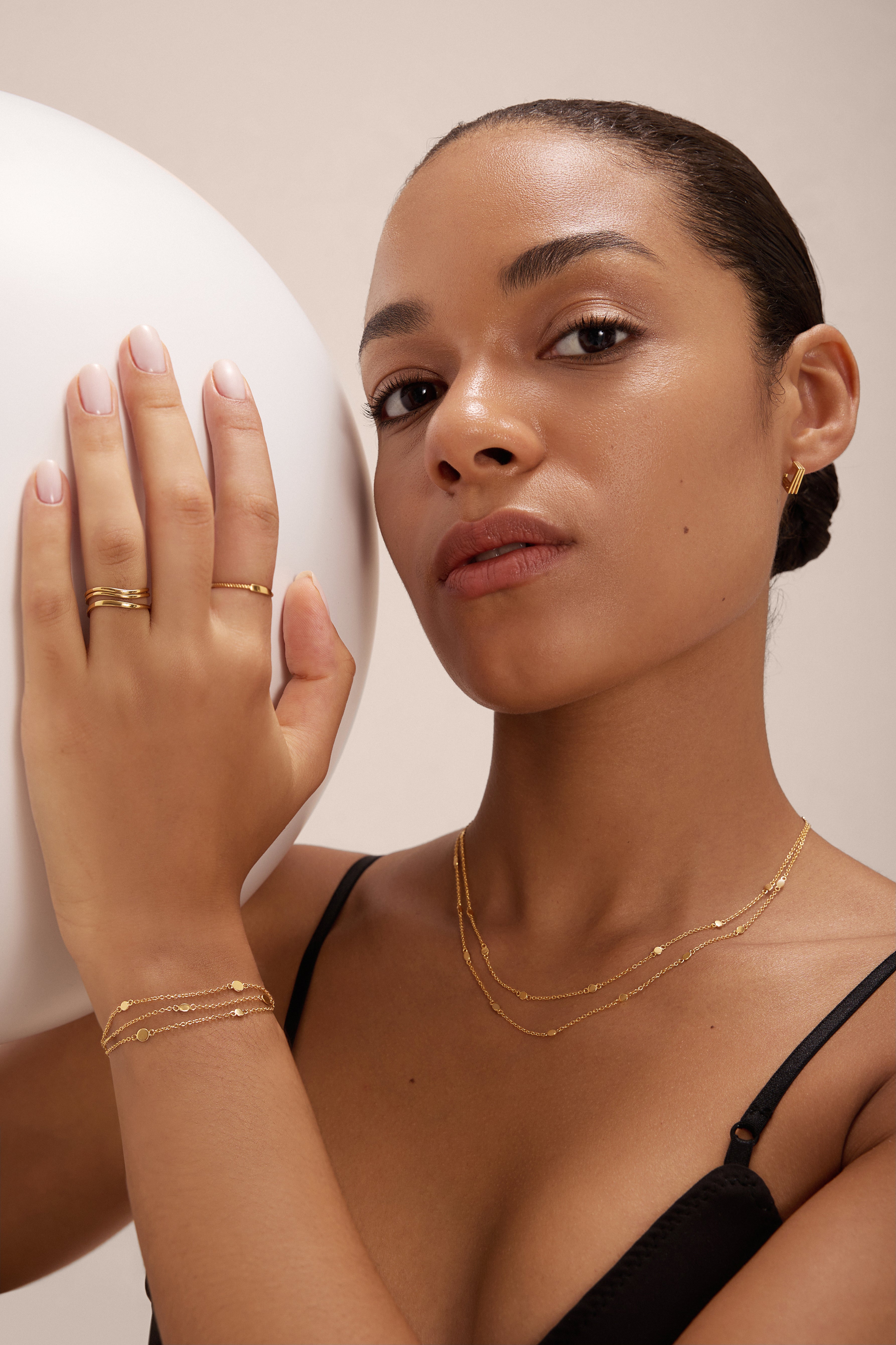 A woman with smooth skin and tied-back hair wears a black top, gold rings, small hoop earrings, and the Simone Triple Chain Layered Bracelet in Gold. She holds a large white sphere in her left hand.