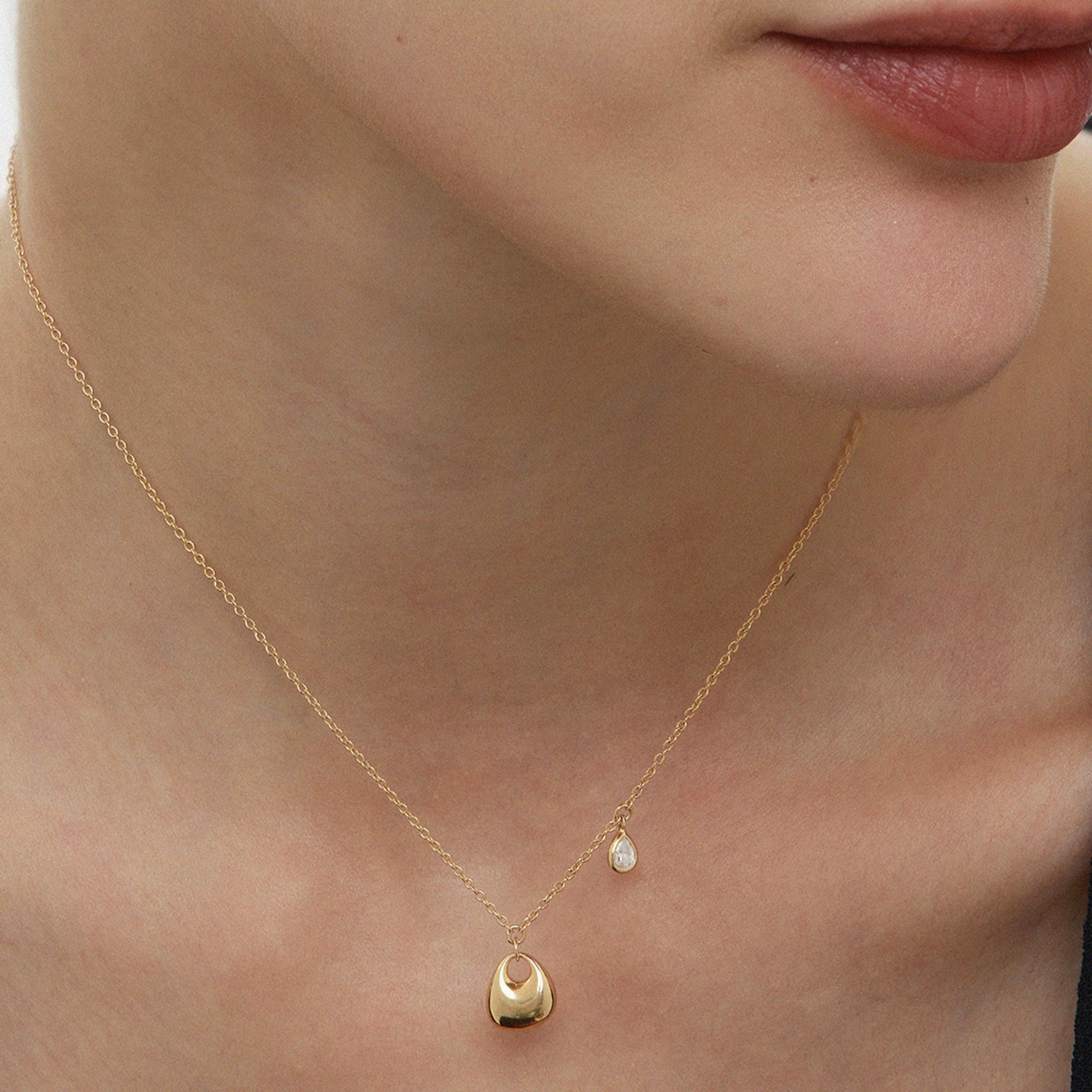 Close-up of a womans neck wearing the Nora Double Pendant Necklace in Gold, featuring a small sculptural gold oval with a circular detail and a tiny teardrop-shaped pearl.