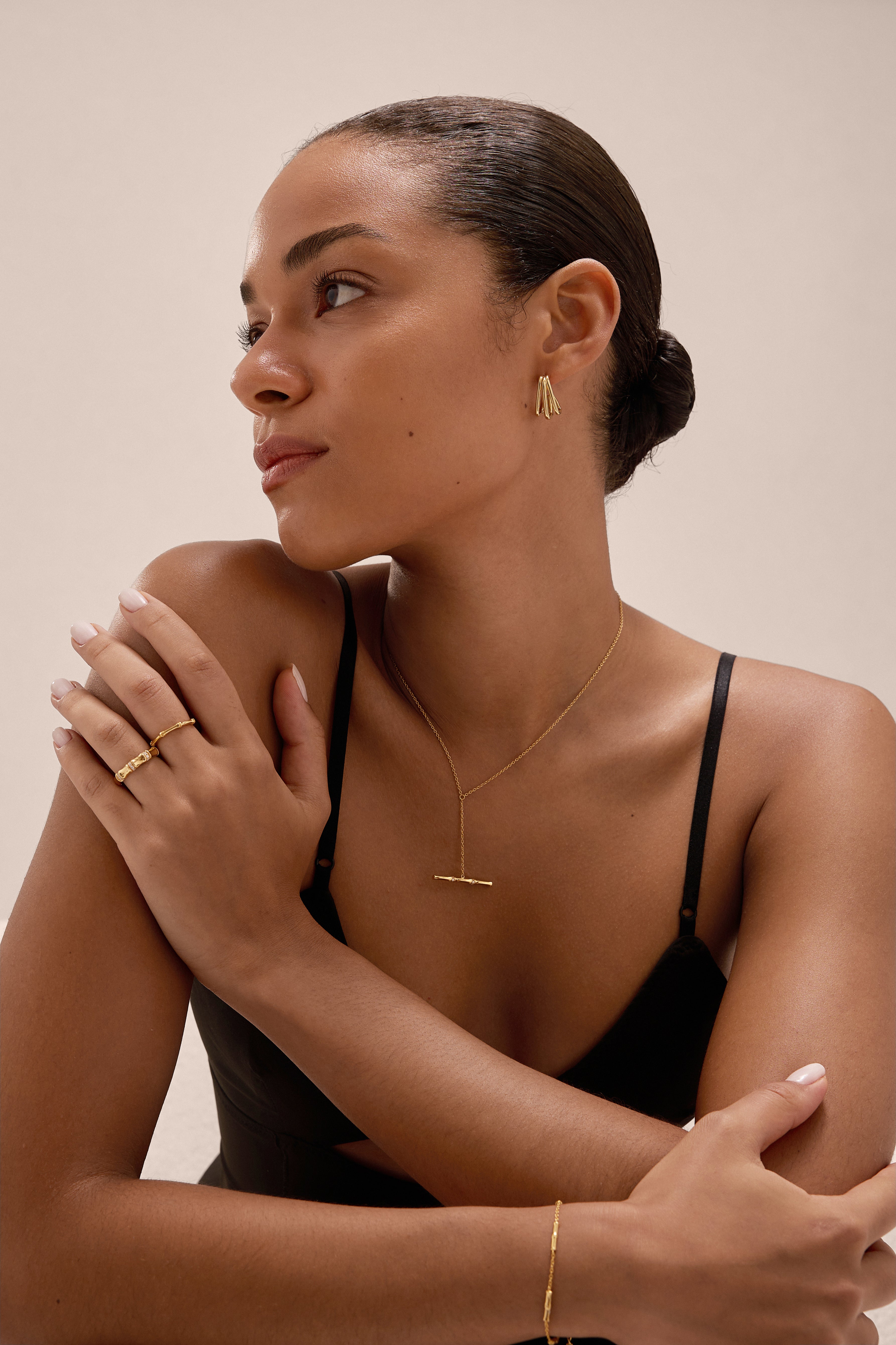 A woman with smooth skin and a neat bun models gold jewelry—earrings, necklace, rings, and the Bamboo Chain Bracelet in Gold. She wears a black spaghetti strap top and looks to the side with one hand on her shoulder.