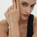 A woman with dark hair pulled back poses thoughtfully in a black sleeveless top, wearing a gold ring and two Bamboo Chain Bracelets in Gold, each featuring delicate 18k gold plating.