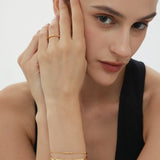 A woman with dark hair pulled back poses thoughtfully in a black sleeveless top, wearing a gold ring and two Bamboo Chain Bracelets in Gold, each featuring delicate 18k gold plating.