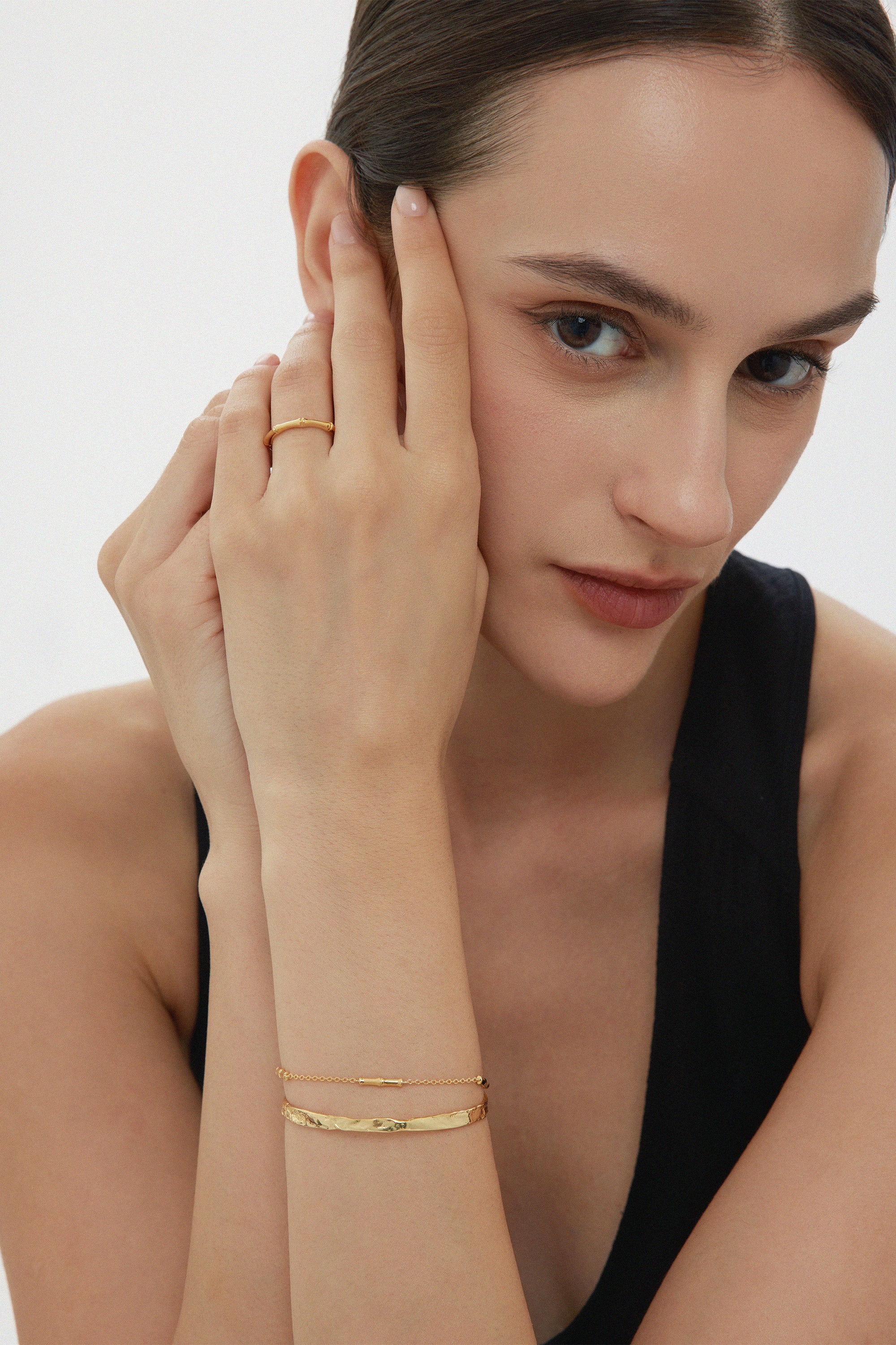 A woman with dark hair pulled back poses thoughtfully in a black sleeveless top, wearing a gold ring and two Bamboo Chain Bracelets in Gold, each featuring delicate 18k gold plating.