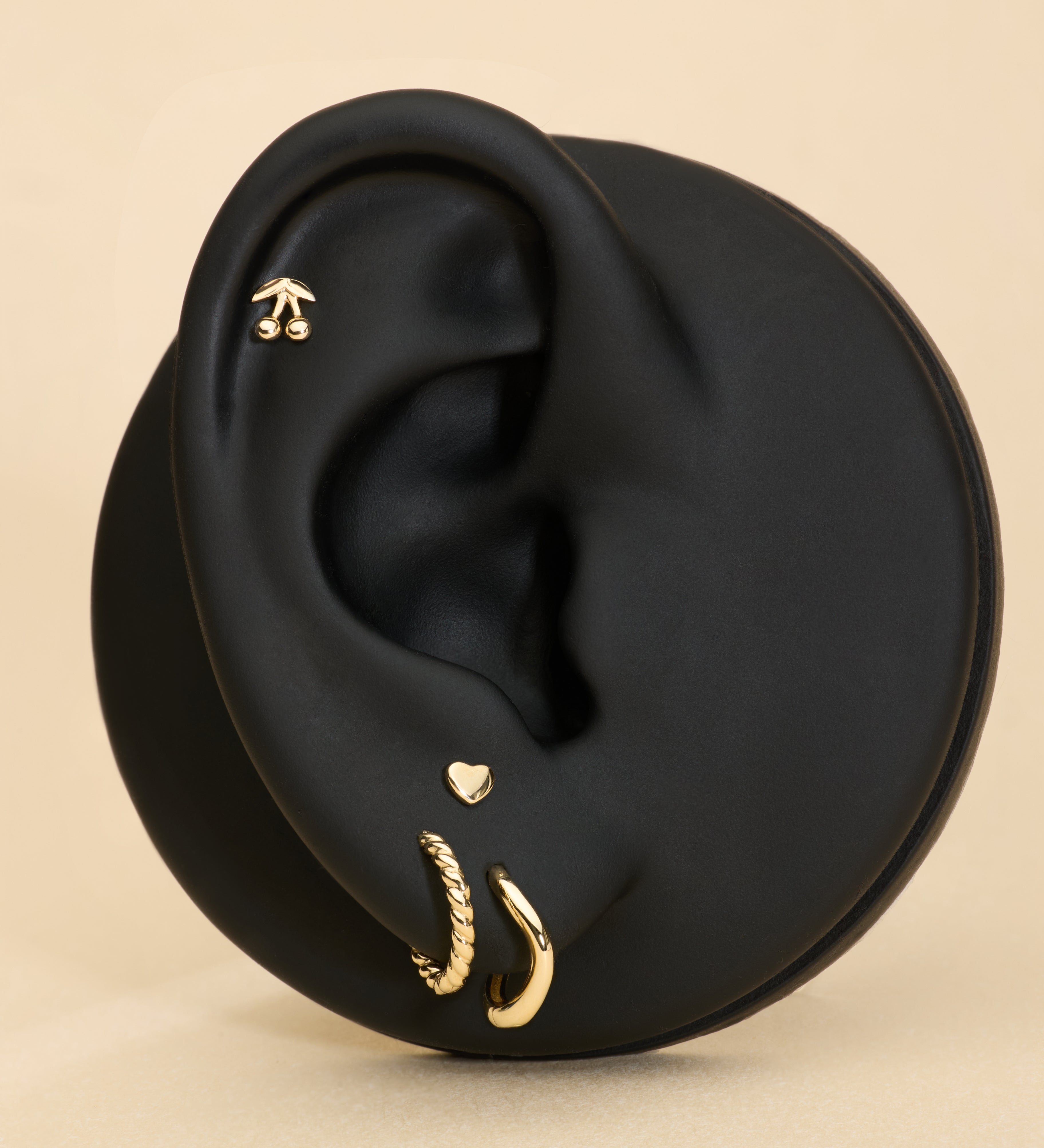 Black ear with gold stud cherry earrings and gold heart flat back stud on a beige background