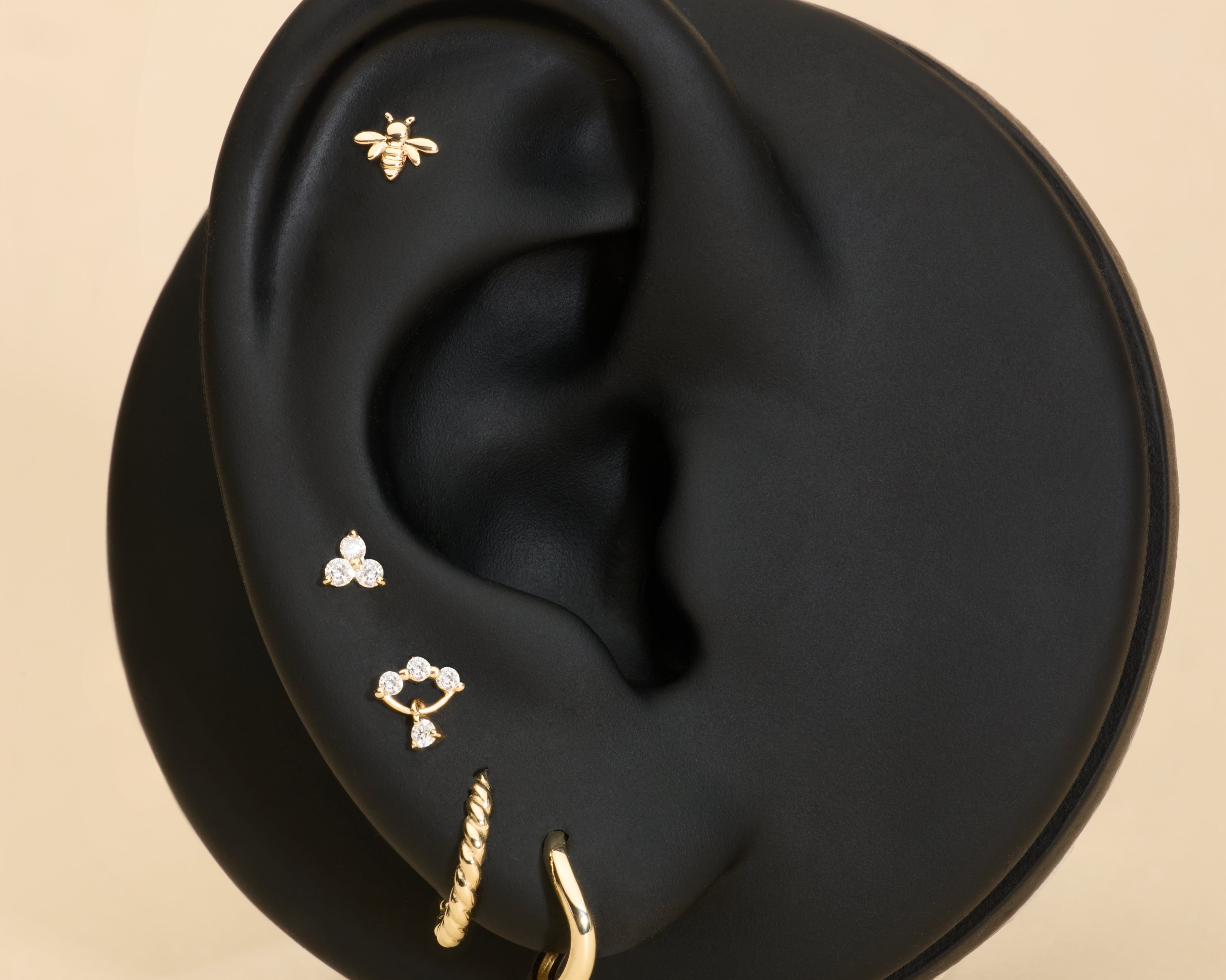 Black ear with gold huggie earrings, bee flatback stud, and diamond flatback stud on a beige background