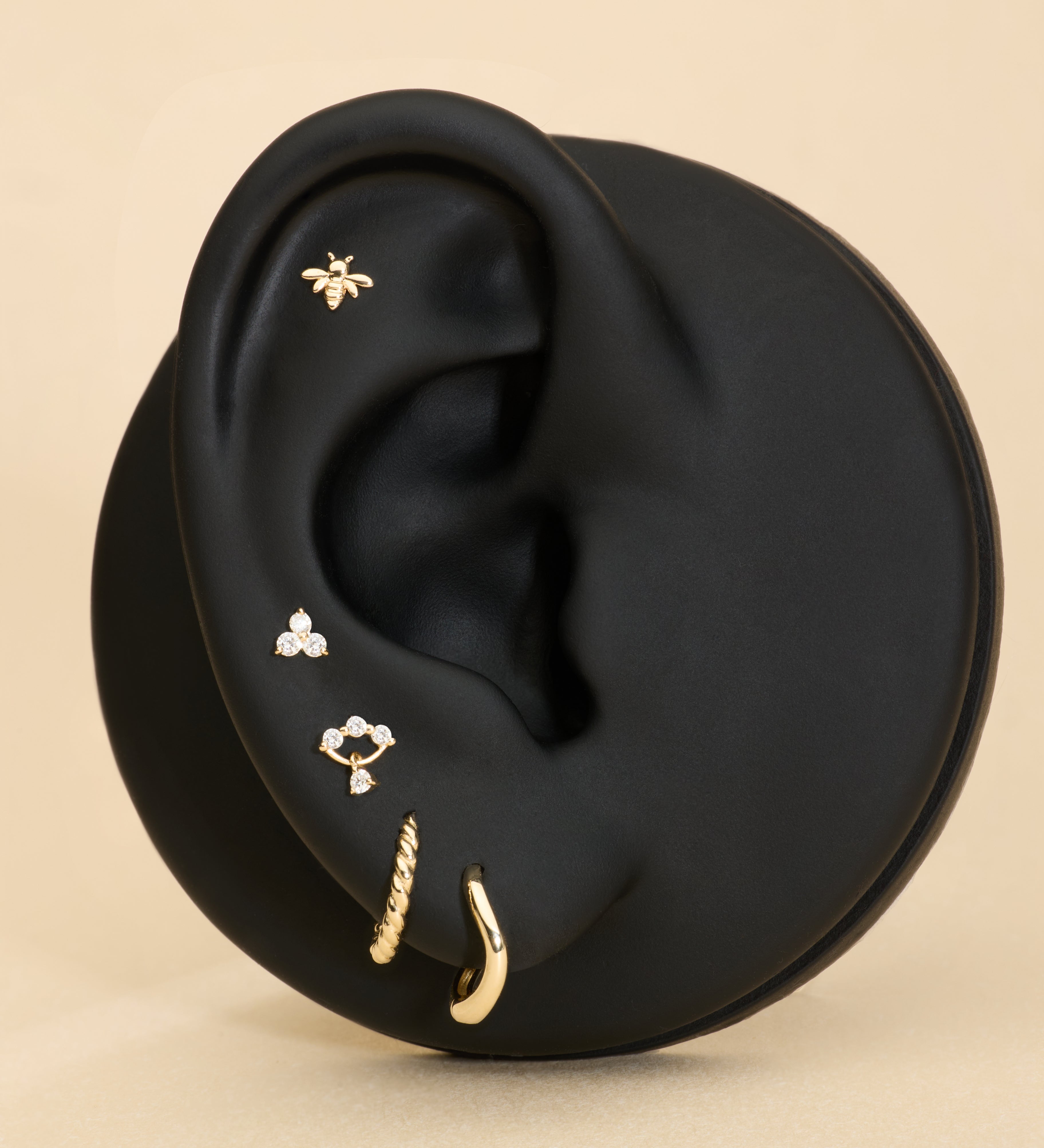 Black ear with gold huggie earrings, bee flatback stud, and diamond flatback stud on a beige background