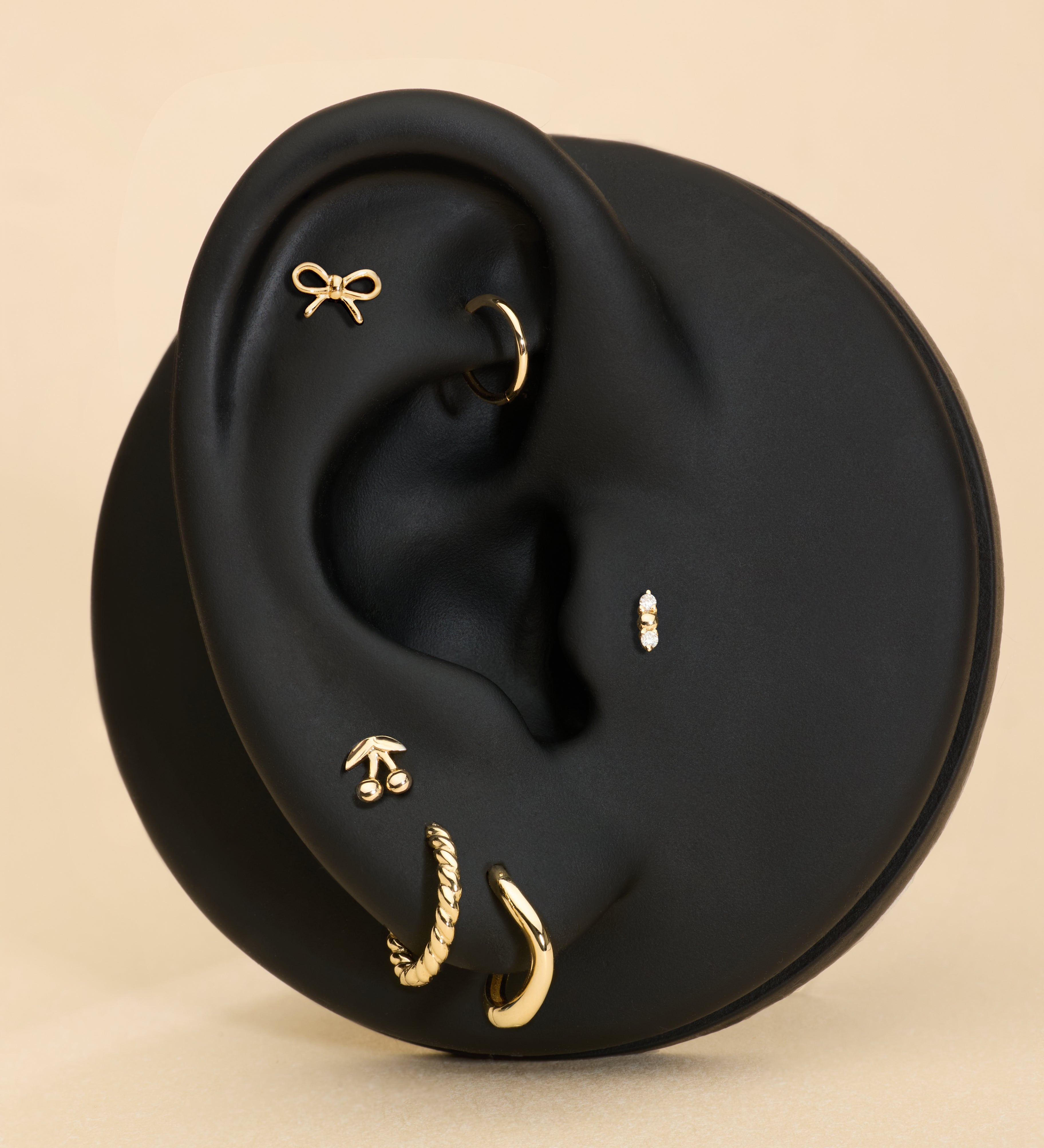 Ear with gold earrings, bow stud, cheery stud and diamond stud on a beige background