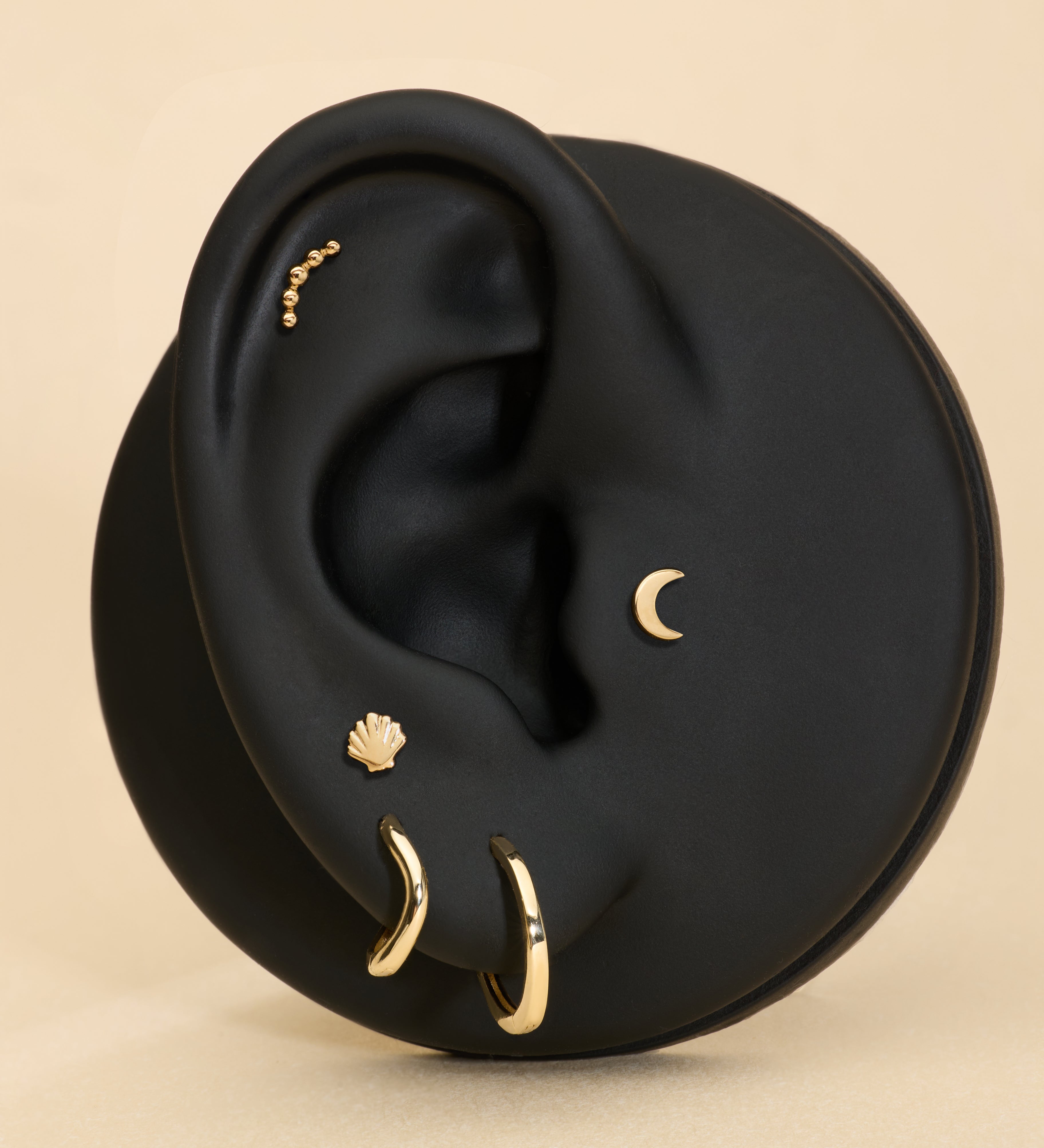 A black ear model showcases the 14K Recycled Solid Yellow Gold Plain Huggie Earring and other sustainable jewelry, set against a beige background.