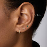 Close-up of a person's ear adorned with stunning piercings: a gold hoop, leaf-shaped stud, green gemstone stud, and the 14K Solid Yellow Gold 20G Mini Clicker in the upper cartilage. The person has short dark hair and wears a black top.