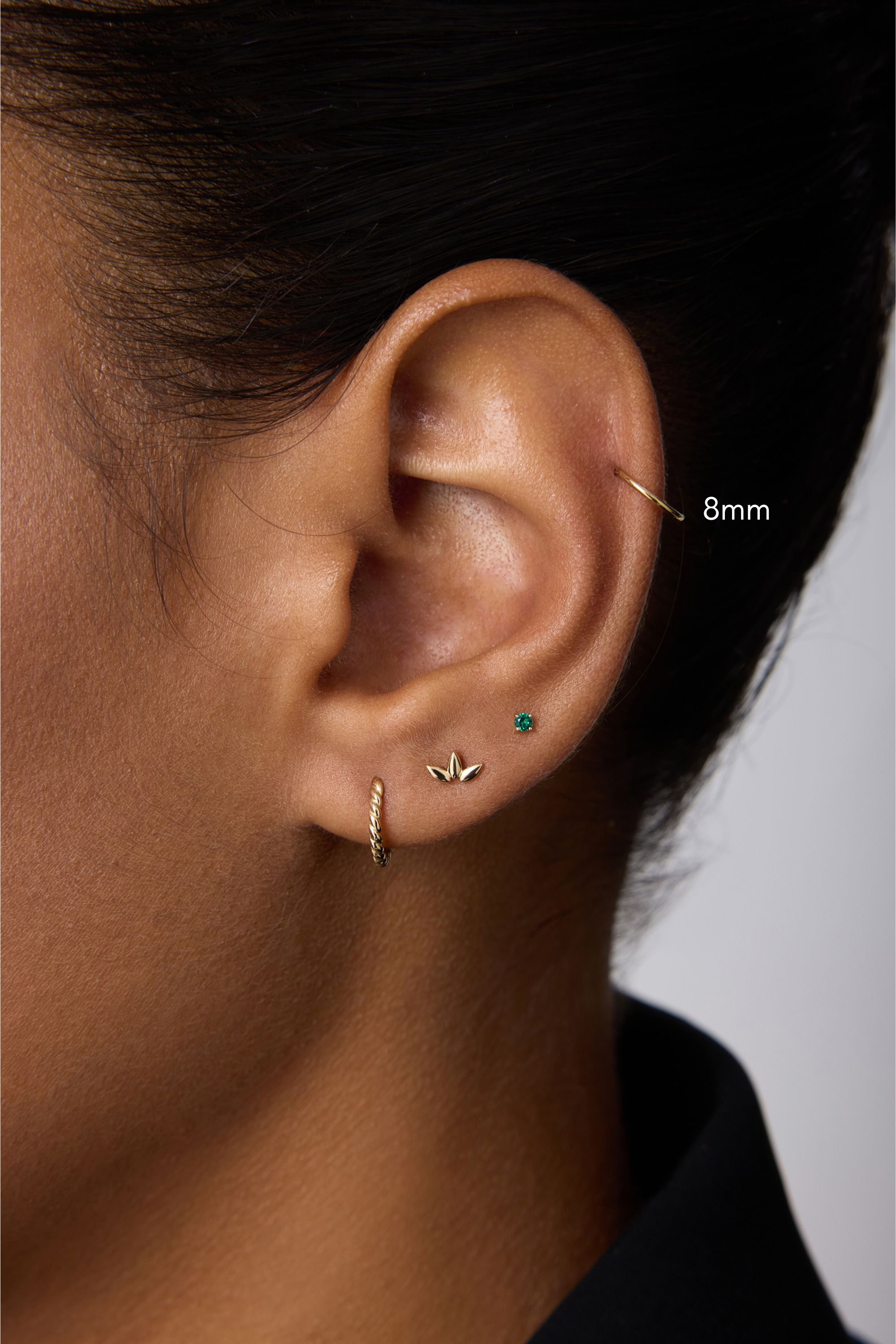 Close-up of a person's ear adorned with stunning piercings: a gold hoop, leaf-shaped stud, green gemstone stud, and the 14K Solid Yellow Gold 20G Mini Clicker in the upper cartilage. The person has short dark hair and wears a black top.