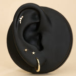 A black mannequin ear showcases the 14K Recycled Solid Yellow Gold Plain Huggie Earring and three other sustainable pieces, all set against a beige background.