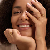 A woman with curly hair smiles, resting her chin on one hand while her other hand, wearing the Aisha Paperclip Stacking Ring in Gold and other rings, partially covers her face. She has light pink nail polish.