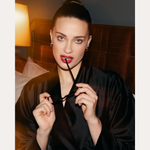 A woman with blue eyes and red lipstick sits on a bed in a dimly lit room, wearing a black robe and Nori Twisted Hoop Earrings in Silver. She holds eyeglasses near her mouth and looks at the camera. Text: SPOTTED ON: GENEVIÈVE CHENNEOUR.
