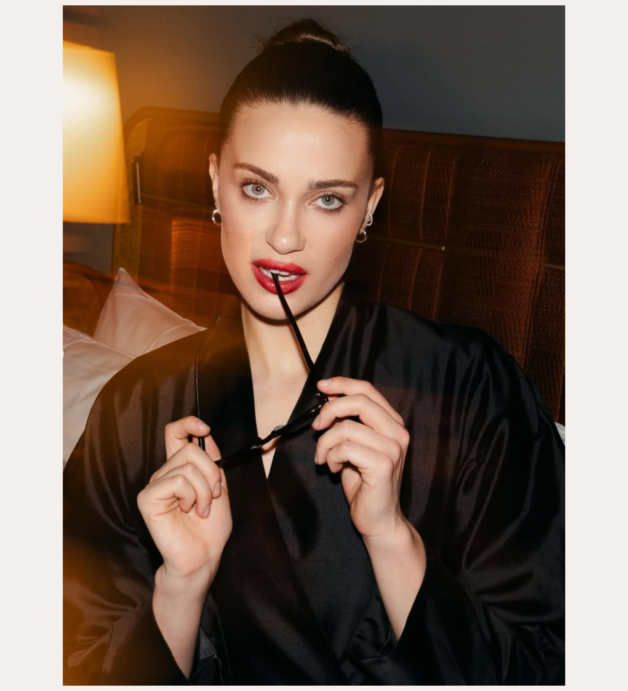 A woman with blue eyes and red lipstick sits on a bed in a dimly lit room, wearing a black robe and Nori Twisted Hoop Earrings in Silver. She holds eyeglasses near her mouth and looks at the camera. Text: SPOTTED ON: GENEVIÈVE CHENNEOUR.