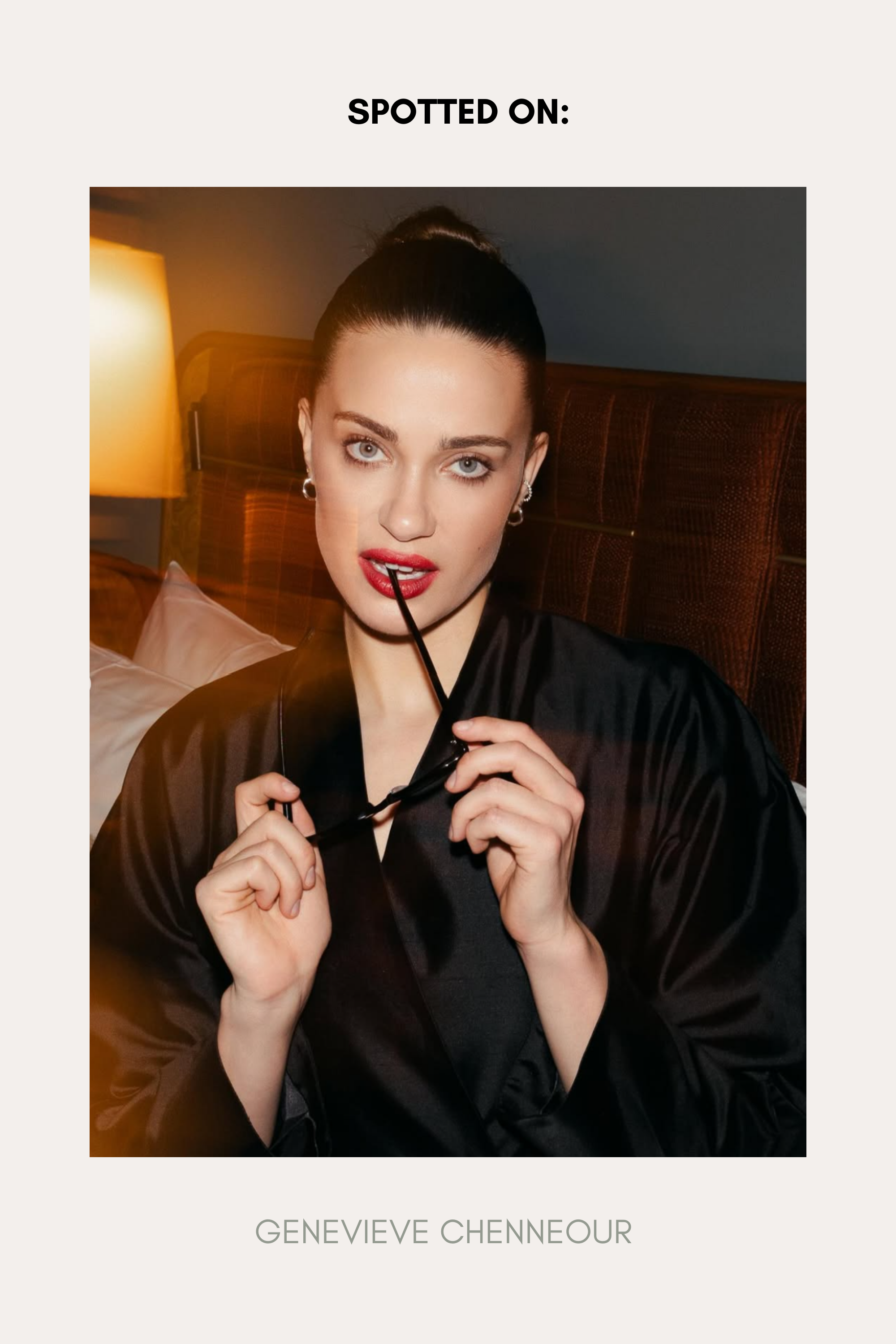 A woman with blue eyes and red lipstick sits on a bed in a dimly lit room, wearing a black robe and Nori Twisted Hoop Earrings in Silver. She holds eyeglasses near her mouth and looks at the camera. Text: SPOTTED ON: GENEVIÈVE CHENNEOUR.