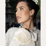 A woman with dark hair in an elegant updo, wearing a textured white outfit with a large flower and bow at the collar, looks to the side. Spotted On: Mimi Webb in Gia Reversible 2 Tone Chubby Hoop Earrings.