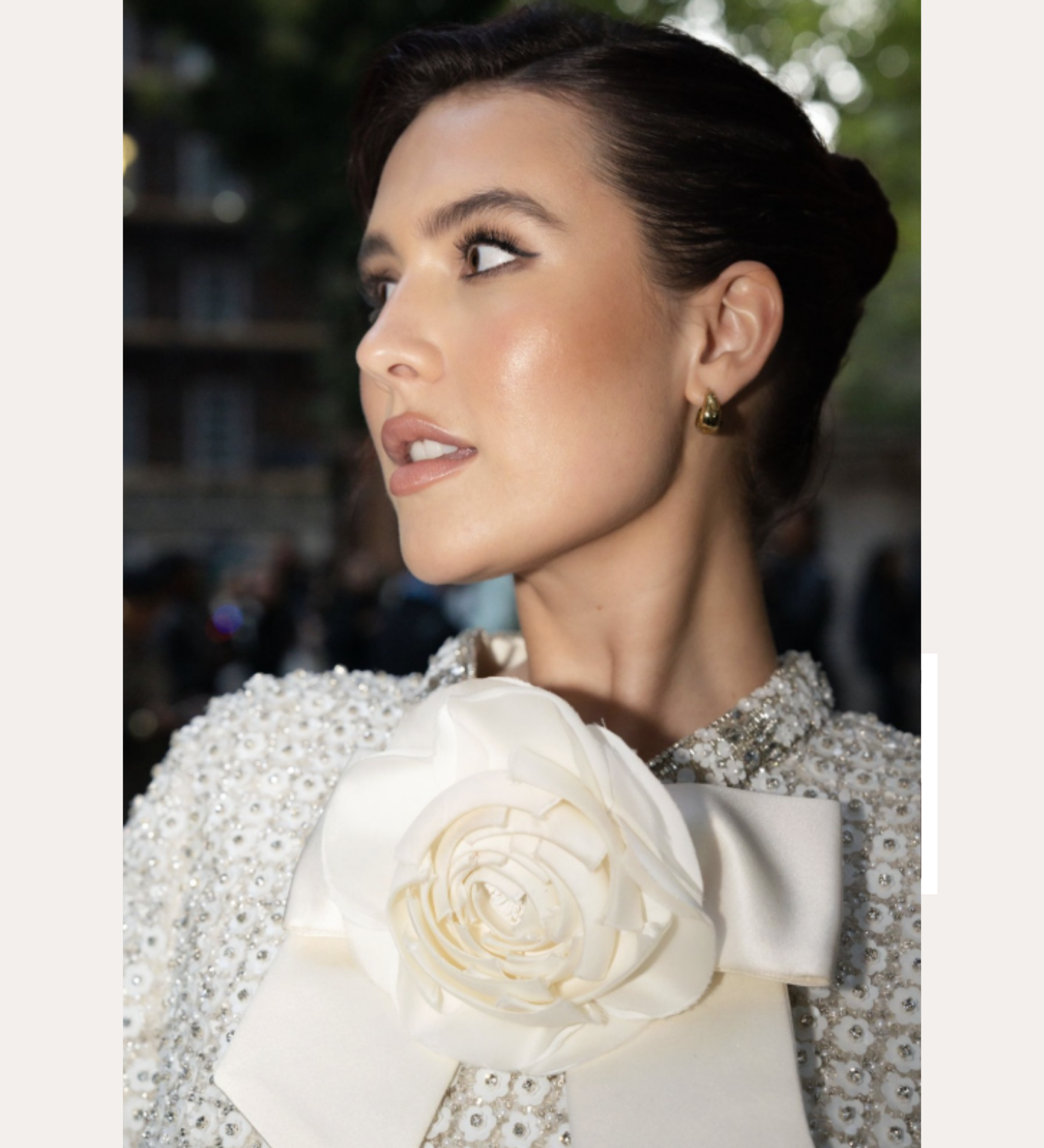 A woman with dark hair in an elegant updo, wearing a textured white outfit with a large flower and bow at the collar, looks to the side. Spotted On: Mimi Webb in Gia Reversible 2 Tone Chubby Hoop Earrings.