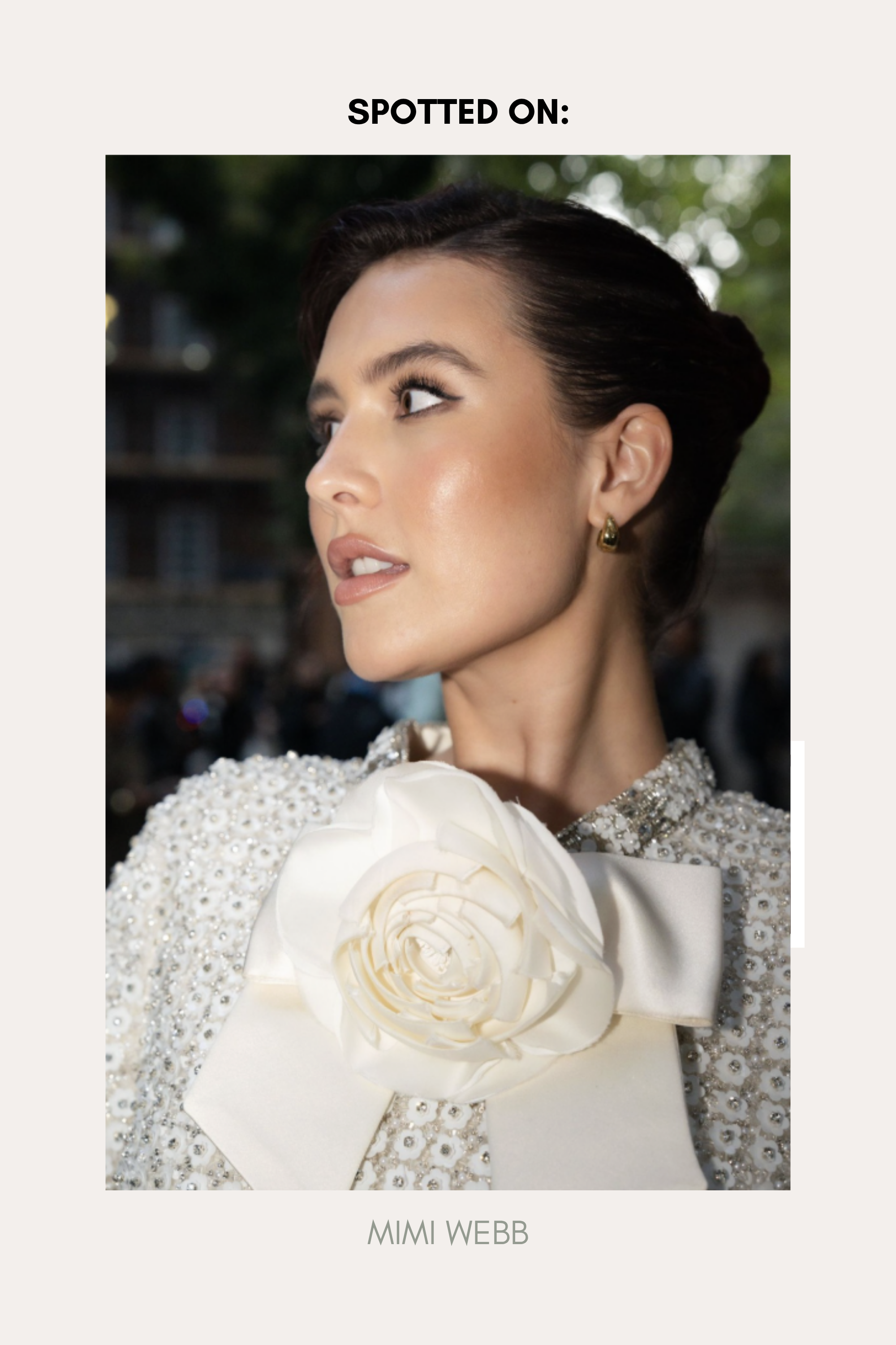 A woman with dark hair in an elegant updo, wearing a textured white outfit with a large flower and bow at the collar, looks to the side. Spotted On: Mimi Webb in Gia Reversible 2 Tone Chubby Hoop Earrings.