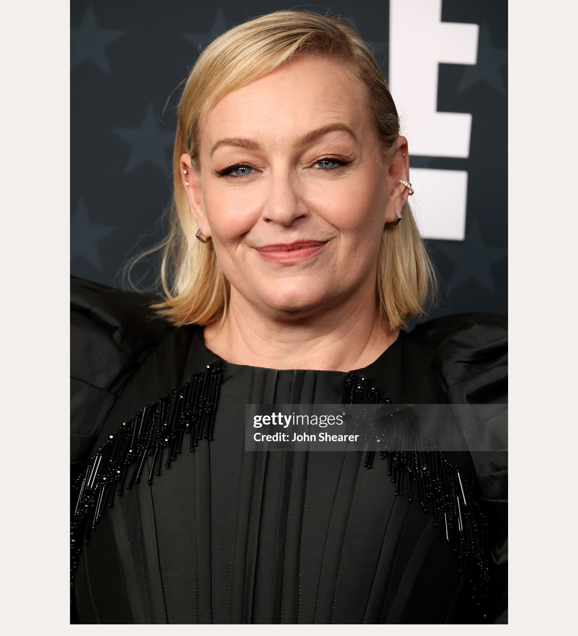 A woman with shoulder-length blonde hair smiles at an event, wearing a black dress and the Judith Hexagon Angular Huggies in Silver. She stands before a gray background with bold white letters. Text reads: Spotted on: Christine Tremarco.