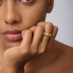 A close-up of a woman with smooth, brown skin resting her chin on her hand, wearing the Bamboo Ring in Gold on her index finger as she gazes calmly past the camera.