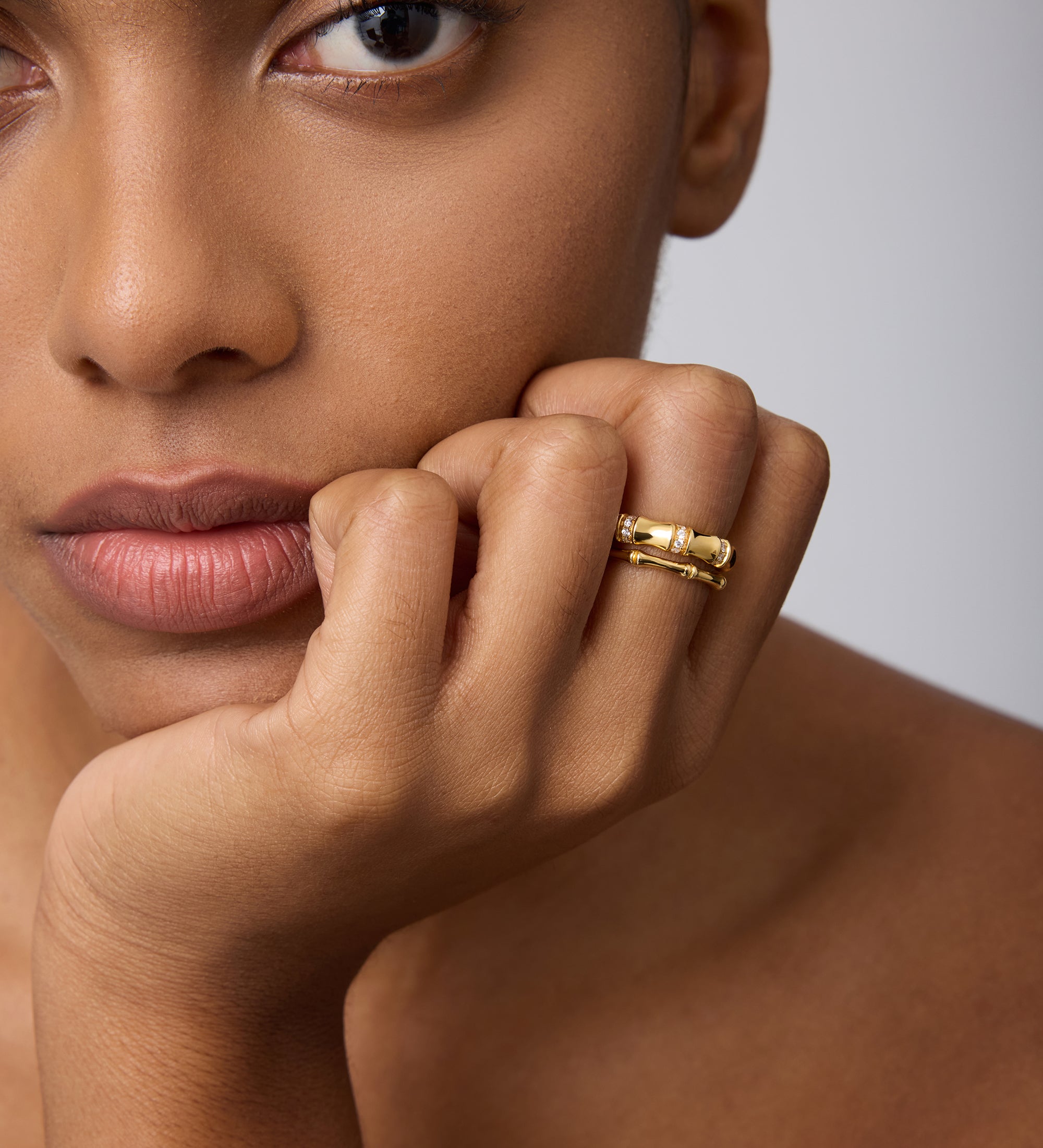 A close-up of a woman with smooth, brown skin resting her chin on her hand, wearing the Bamboo Ring in Gold on her index finger as she gazes calmly past the camera.