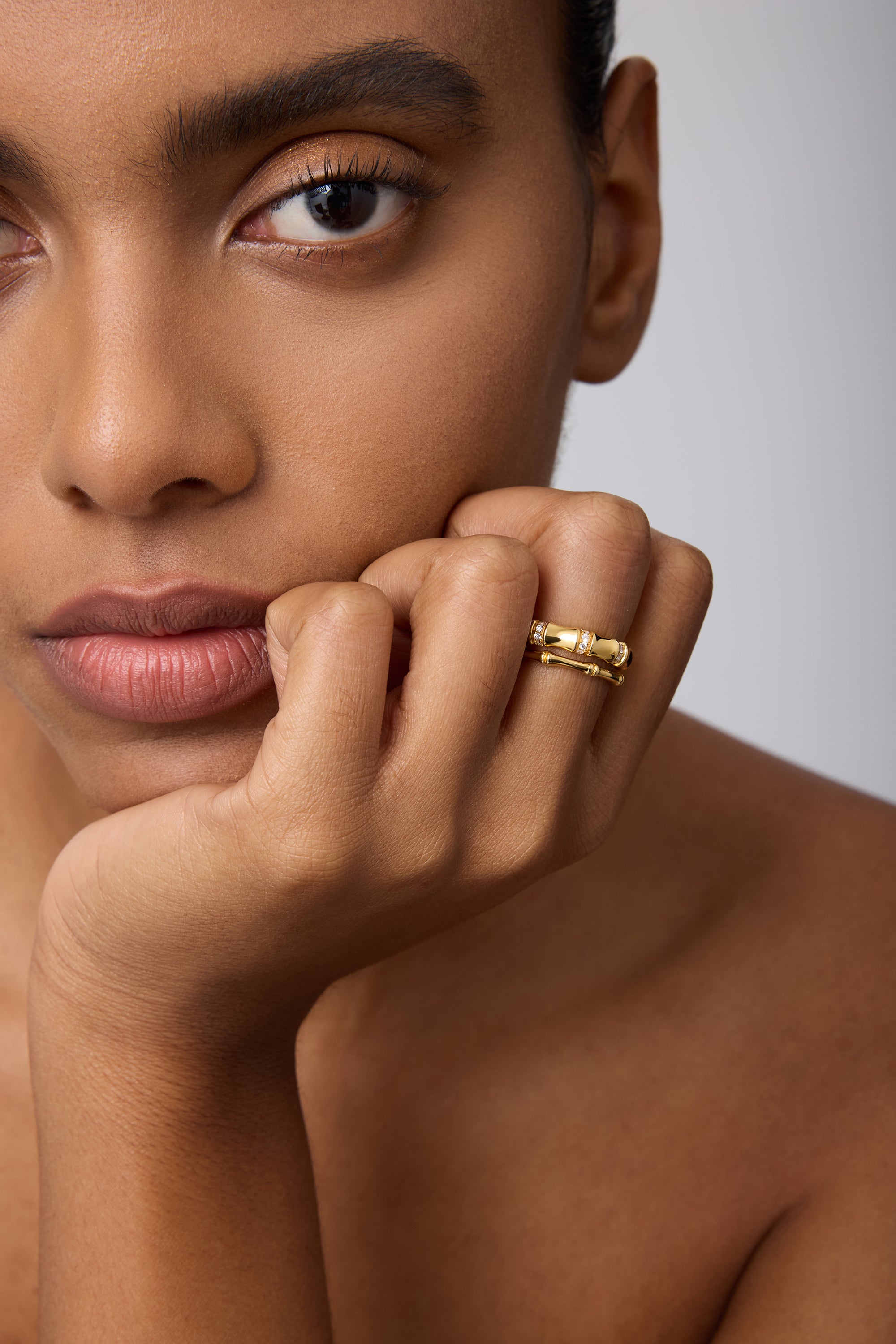 A close-up of a woman with smooth, brown skin resting her chin on her hand, wearing the Bamboo Ring in Gold on her index finger as she gazes calmly past the camera.