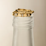 The Bamboo Ring in Gold sits elegantly on the frosted neck of a glass bottle, accented by a pair of gold hoop earrings against a neutral background for a minimalist and sophisticated display.